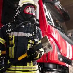 Bombeiro do curso de Pós-graduação em Engenharia de Segurança de Combate a Incêndio do eniac atrás de um caminhão dos bombeiros, utilizando uniforme e segurando um machado